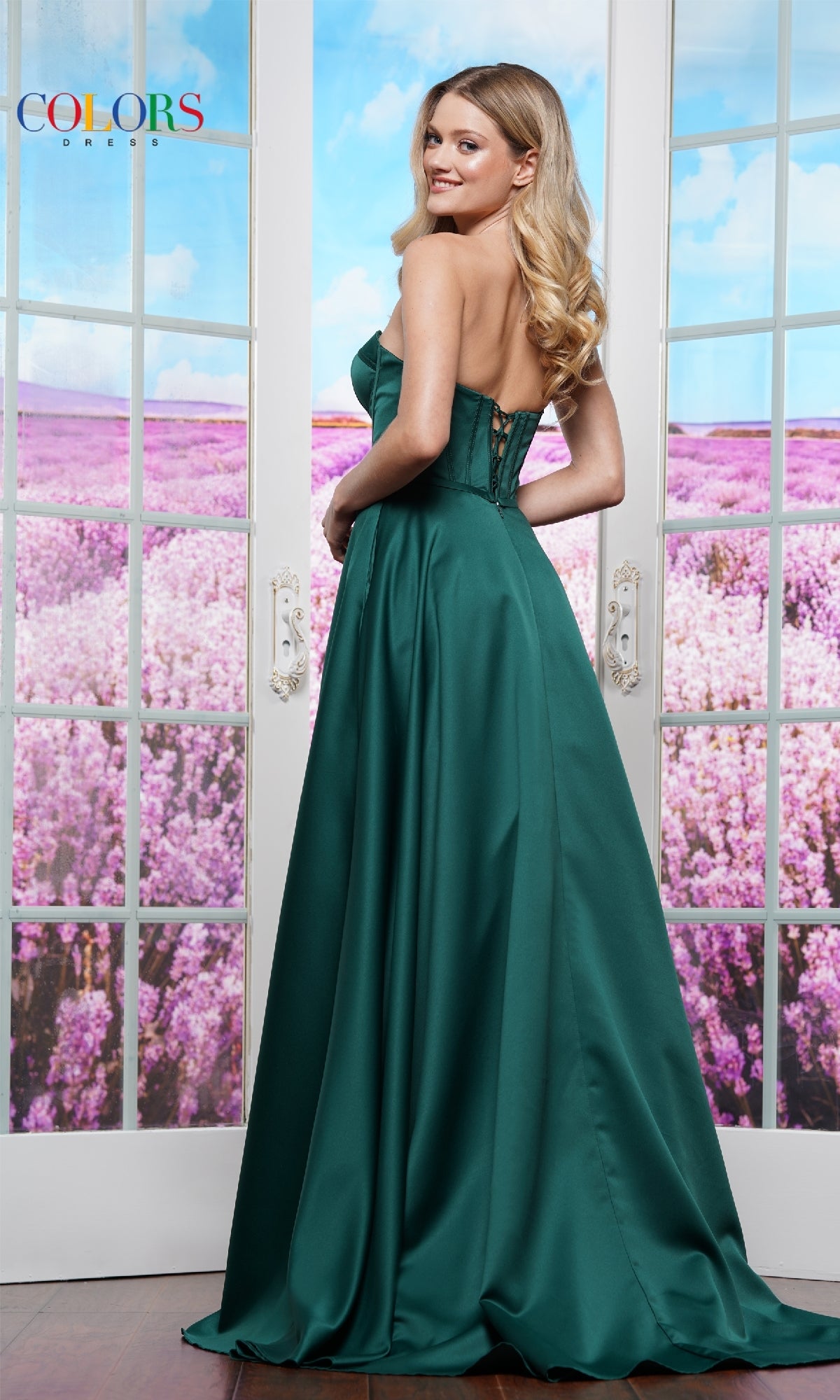 A woman wearing the Strapless Corset Prom Dress: Colors Dress 3434 stands indoors before glass doors that overlook a field of purple flowers.